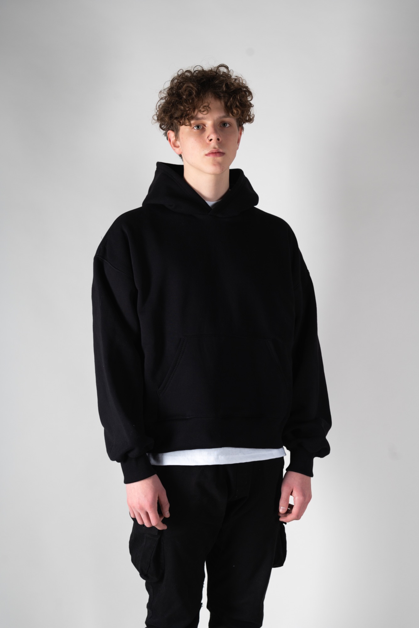 Młody mężczyzna z kręconymi włosami w czarnej, luźnej bluzie z kapturem i czarnych spodniach cargo na jasnoszarym tle.