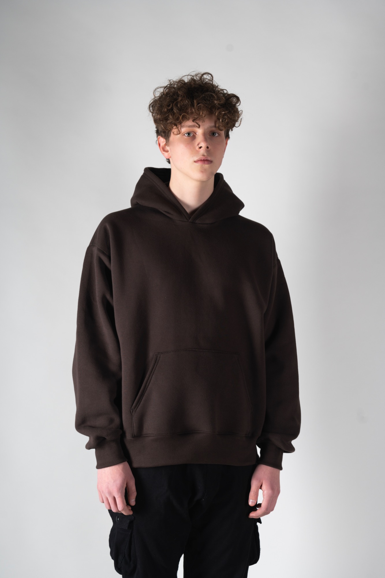Młody mężczyzna z kręconymi włosami prezentuje brązową, luźną bluzę z kapturem na jasnoszarym tle.