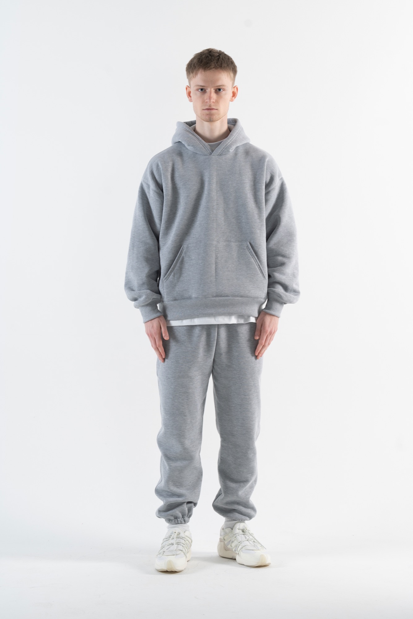 Mężczyzna w szarym dresie z kapturem i białych sneakersach na białym tle studyjnym.