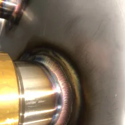 Szczegółowe zbliżenie na połączenie spawane rury ze zbiornikiem, widoczne ślady obróbki na powierzchni zbiornika i charakterystyczne zabarwienie spoiny.