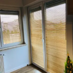 Wnętrze pokoju z oknami i drzwiami balkonowymi osłoniętymi jasnymi żaluzjami poziomymi, obok drzwi stoi mała choinka.