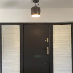 Ciemne drzwi wejściowe flankowane oknami z białymi plisami, oświetlone przez minimalistyczną lampę wiszącą.