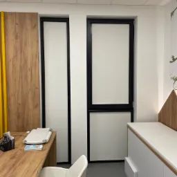 Wnętrze biura z dwoma oknami z białymi roletami i czarnymi ramami, biurko z przyborami, białe krzesło i szafka z siedziskiem.