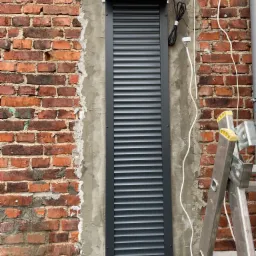 Montaż zewnętrznej osłony okiennej na ceglanej ścianie, widoczne przewody elektryczne i drabina oparta o ścianę.