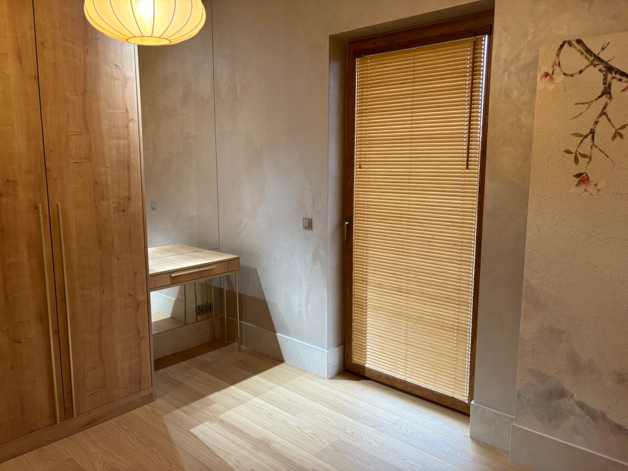 Beżowe żaluzje poziome w drewnianej ramie okna w pokoju z drewnianą podłogą i szafą. Na ścianie dekoracyjny motyw kwiatowy.