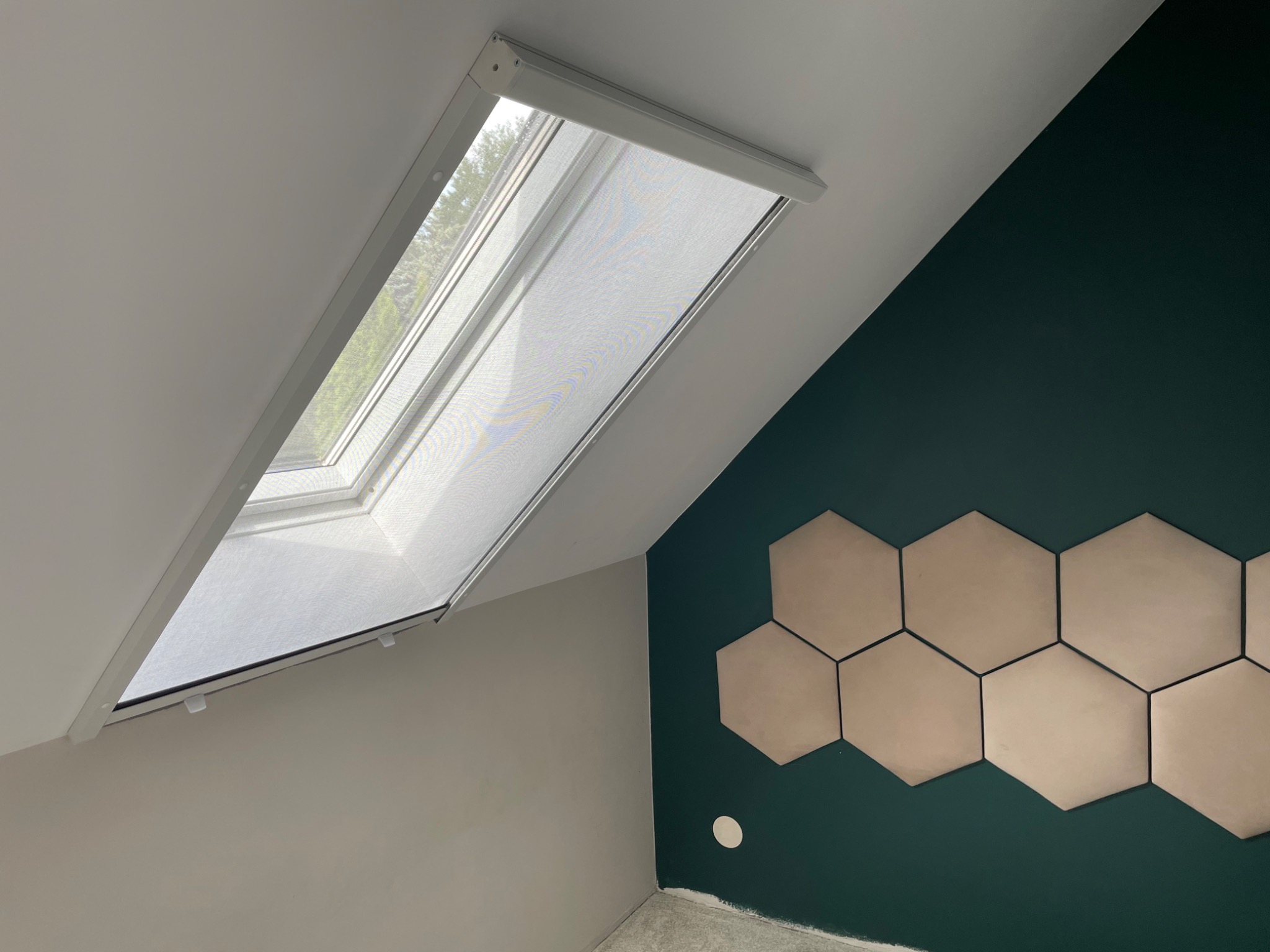 Okno dachowe z zamontowaną siatką przeciw owadom, widok z wnętrza, ściana w kolorze butelkowej zieleni z dekoracyjnymi heksagonami.