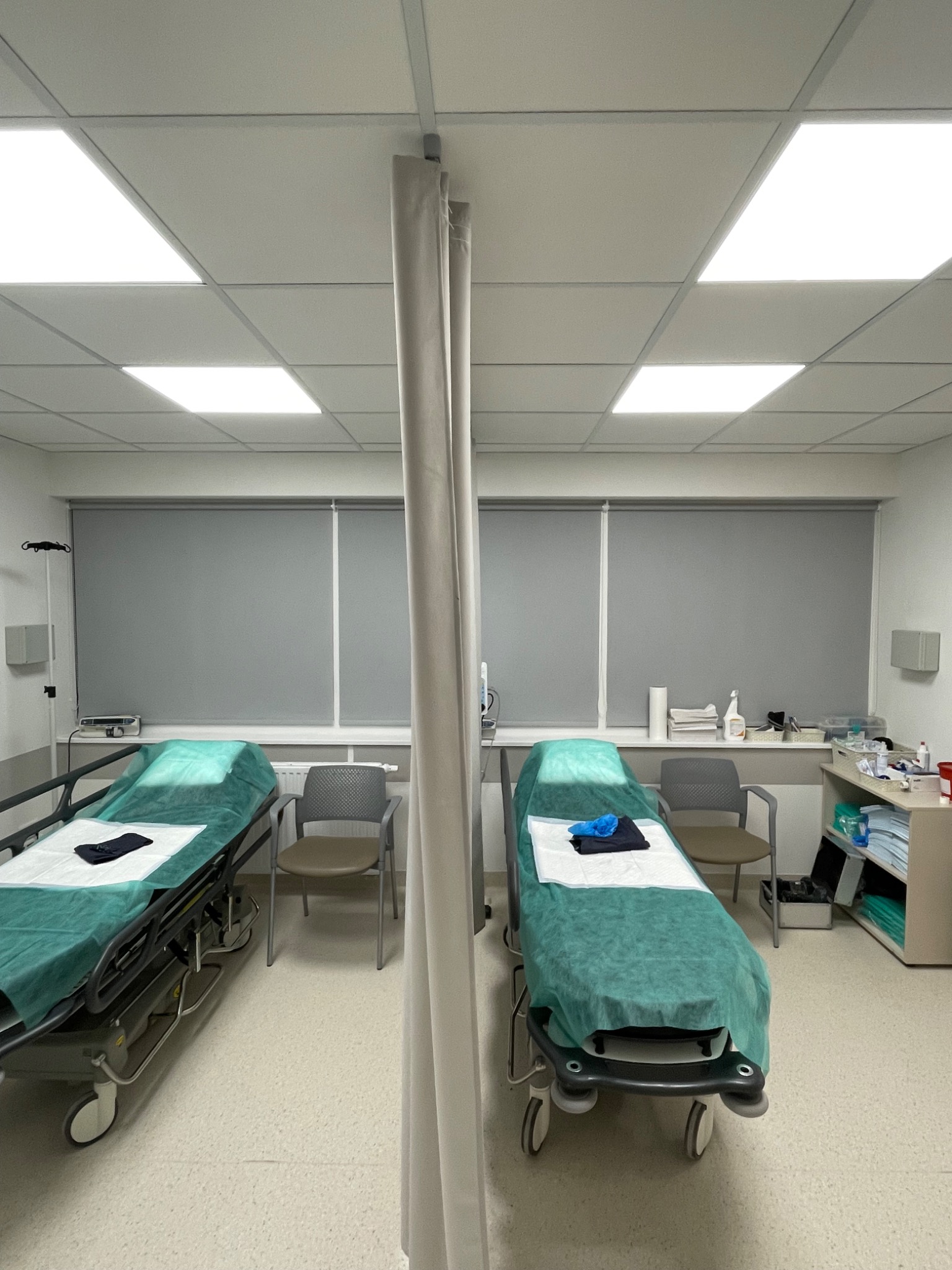 Dwa łóżka szpitalne z zasuniętymi szarymi roletami w oknach, widok z góry.