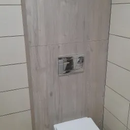 Wisząca toaleta z białym siedziskiem, ukryta spłuczka z chromowanym panelem na ścianie wykończonej panelami imitującymi drewno, obok płytki w odcieniach beżu, w rogu widoczne środki czystości.