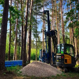 Wiertnica Volvo EC18E na tle lasu, obok sterta żwiru i rury PCV. Maszyna gotowa do wiercenia, widoczna w leśnym otoczeniu.
