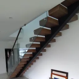 Drewniane schody wspornikowe z metalową konstrukcją i szklaną balustradą, mocowaną punktowo, w jasnym wnętrzu z marmurową podłogą i obrazami w ramkach przy ścianie.