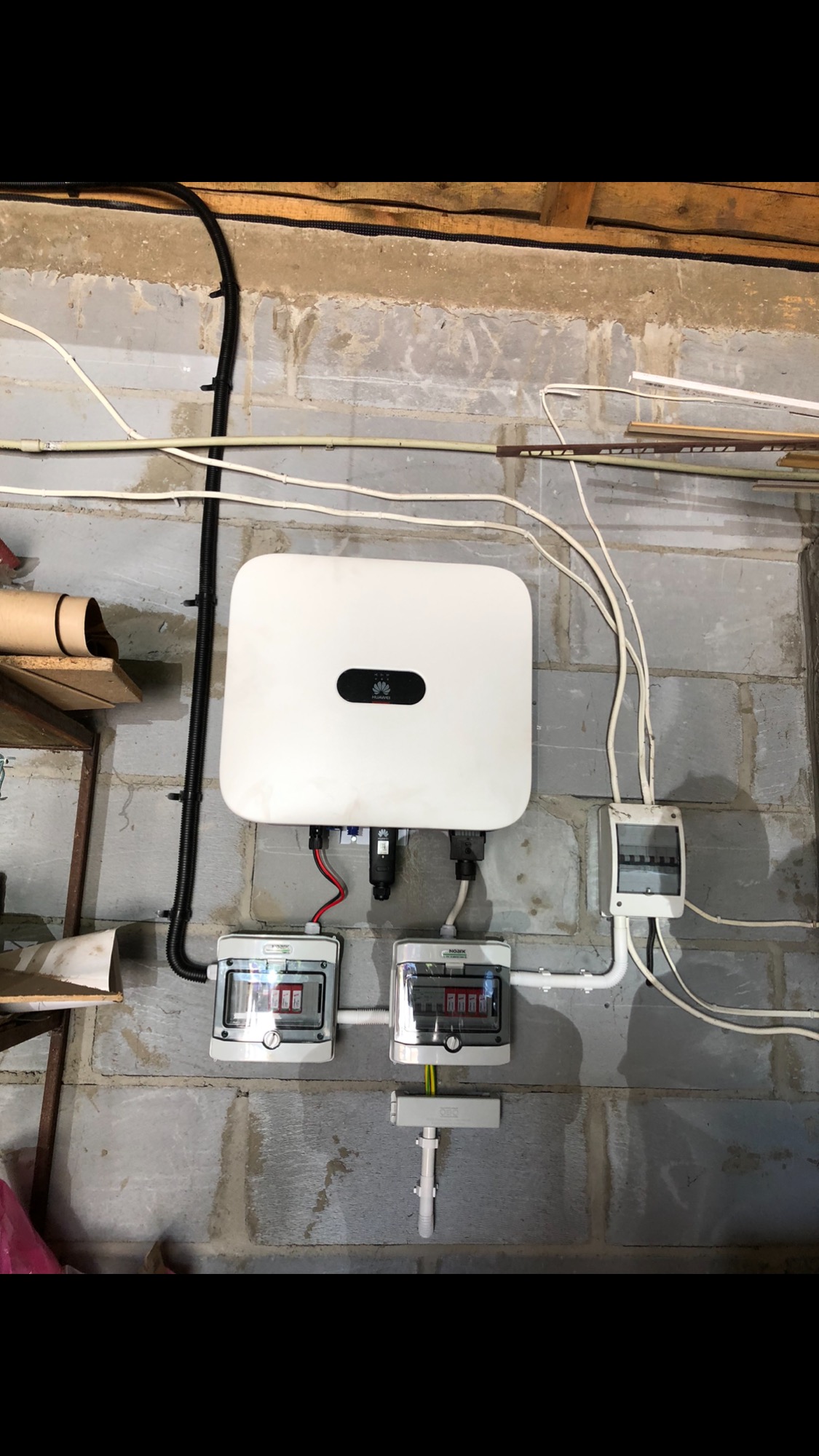 Biały falownik Huawei zamontowany na szarej ścianie z bloków betonowych, z widocznymi przewodami, dwoma rozdzielnicami i czarną rurą karbowaną.