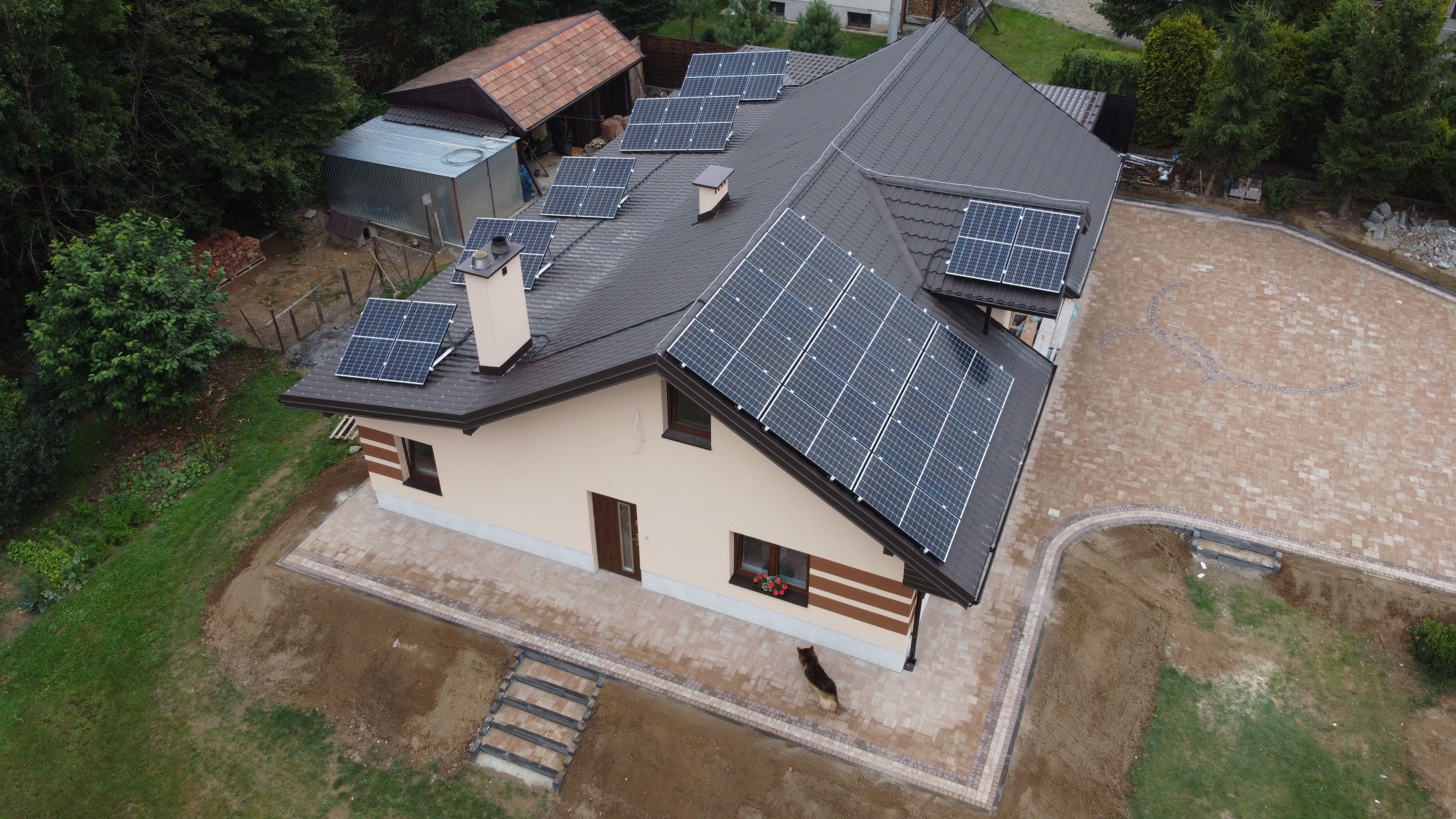 Dom jednorodzinny z brązowym dachem pokrytym panelami słonecznymi, widok z góry, na podwórku widoczny pies rasy owczarek niemiecki.