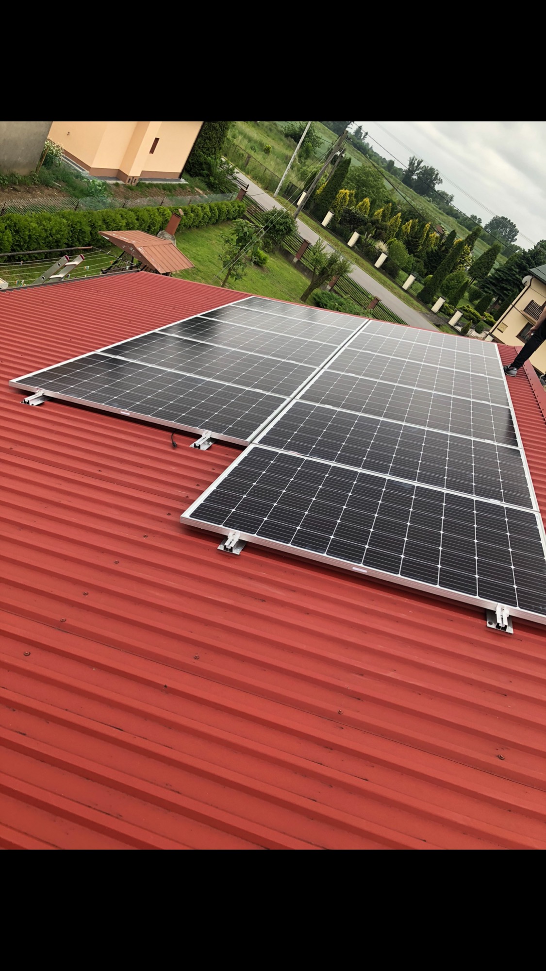 Panele słoneczne zamontowane na czerwonym, blaszanym dachu domu jednorodzinnego, widok z góry na instalację i okoliczną zabudowę.