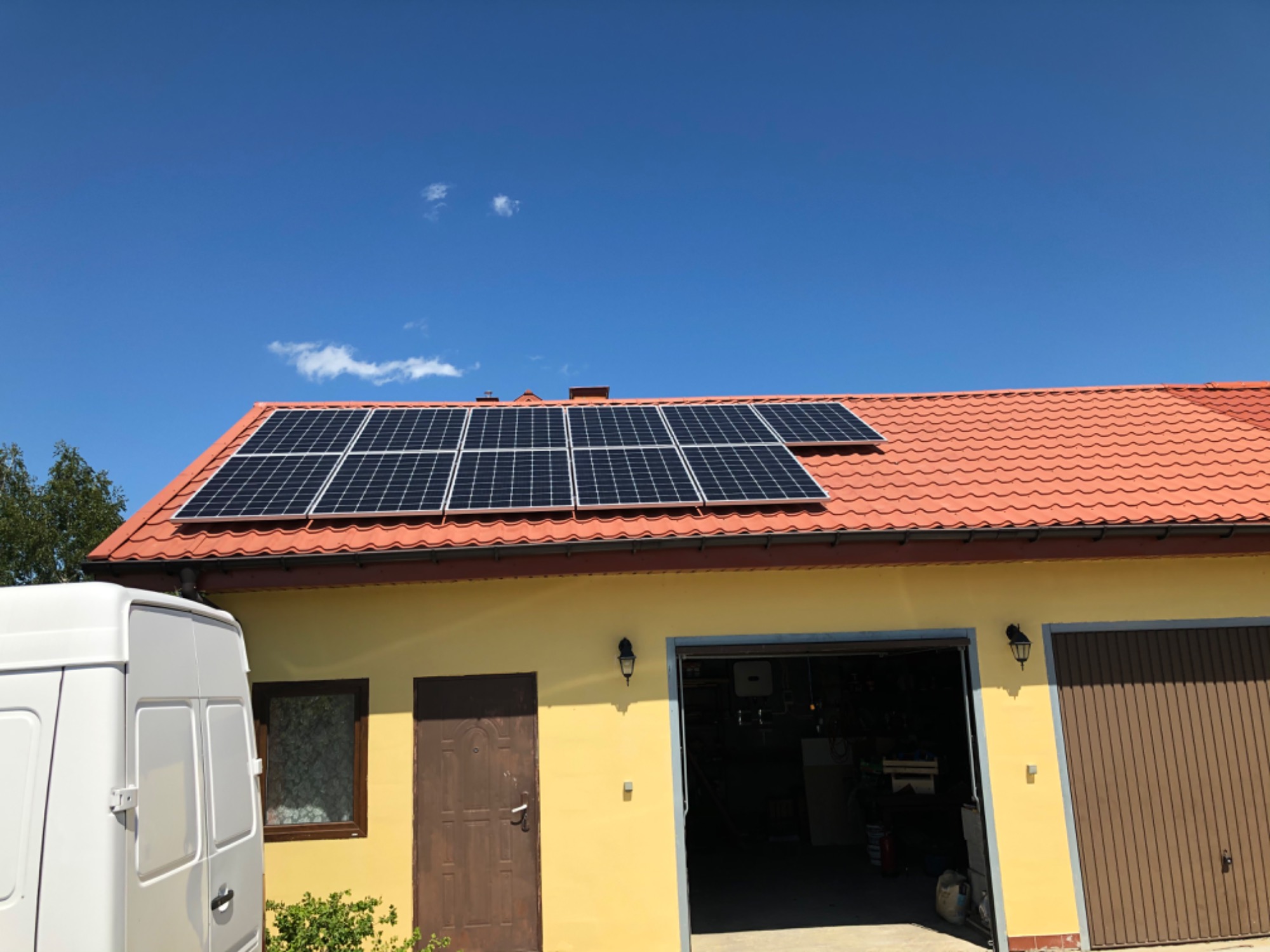 Panele słoneczne na dachu garażu z czerwoną dachówką, biały samochód dostawczy zaparkowany obok.