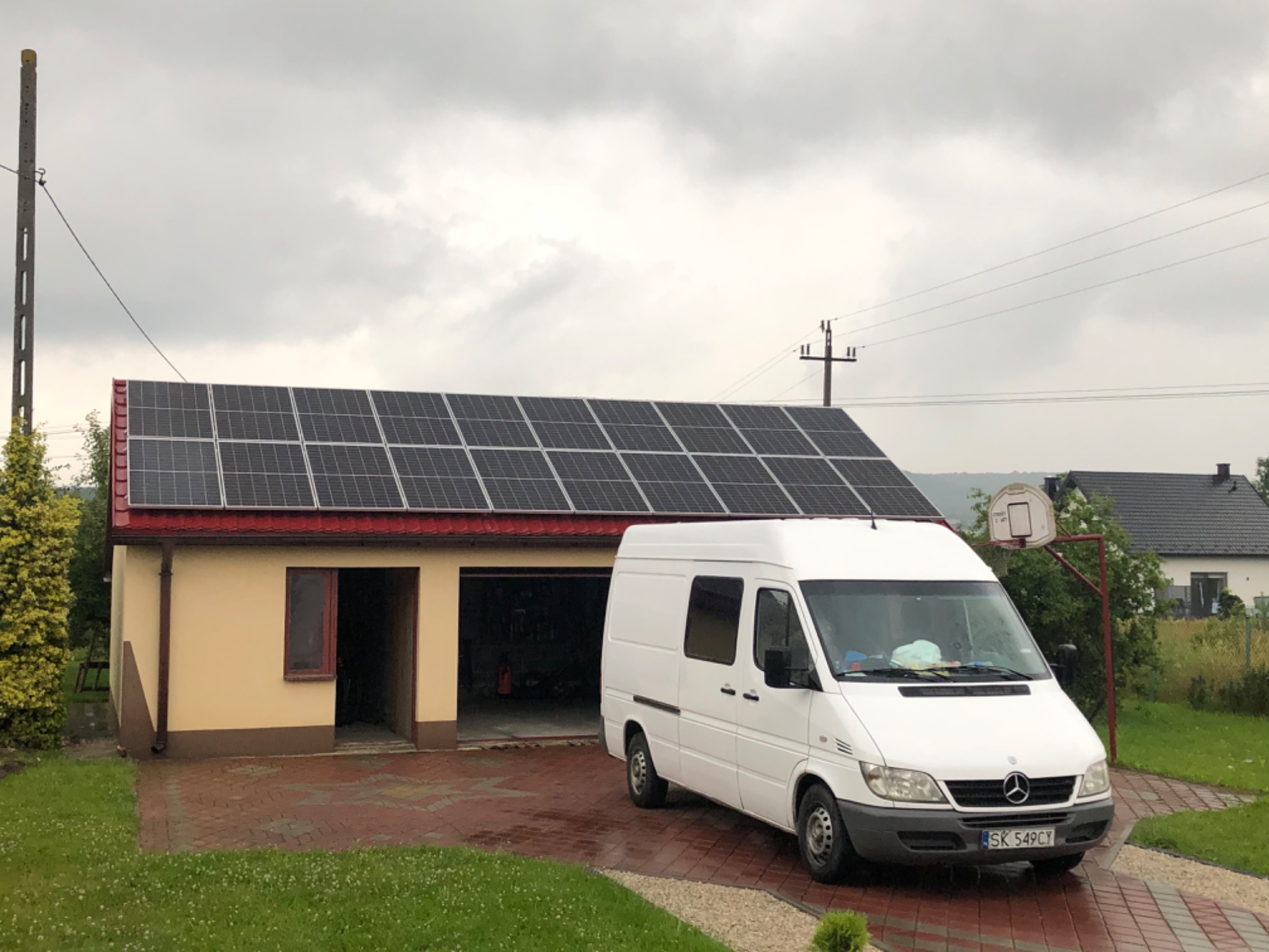 Instalacja paneli słonecznych na dachu garażu z czerwonymi obróbkami blacharskimi, na podjeździe stoi biały samochód dostawczy Mercedes Sprinter, w tle dom z koszem do koszykówki.