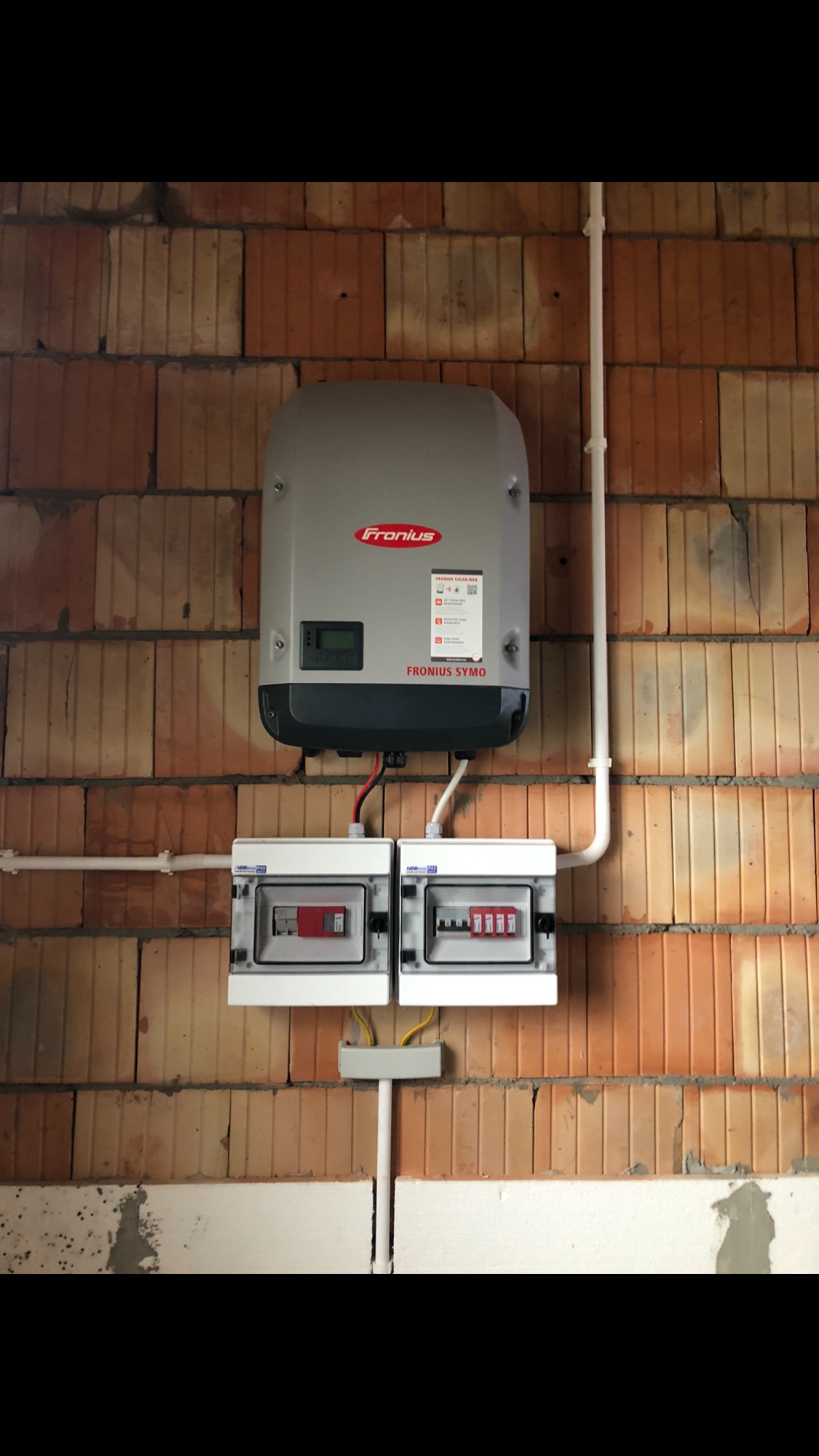 Zamontowany inwerter Fronius Symo oraz dwie skrzynki bezpiecznikowe na ceglanej ścianie, instalacja fotowoltaiczna w trakcie podłączania.