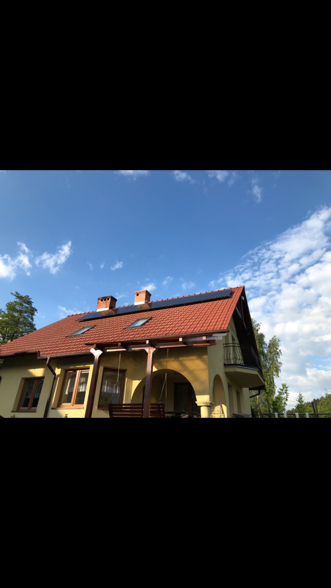 Żółty dom jednorodzinny z czerwoną dachówką i panelami słonecznymi na dachu, widoczne kominy, okna dachowe i balkon, na tle błękitne niebo z chmurami.