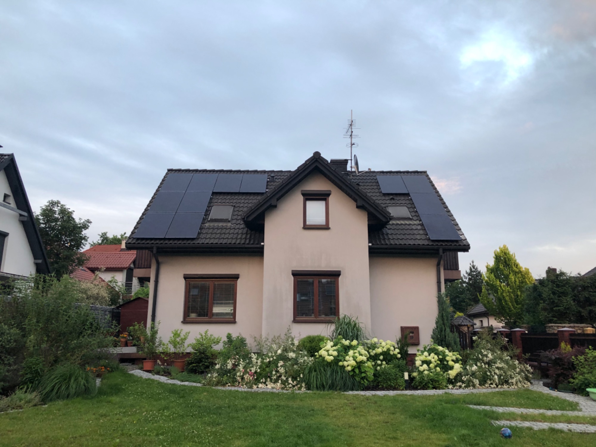 Dom jednorodzinny z beżową elewacją i panelami słonecznymi na dachu, widok z przodu na zadbany ogród z kwitnącymi hortensjami.