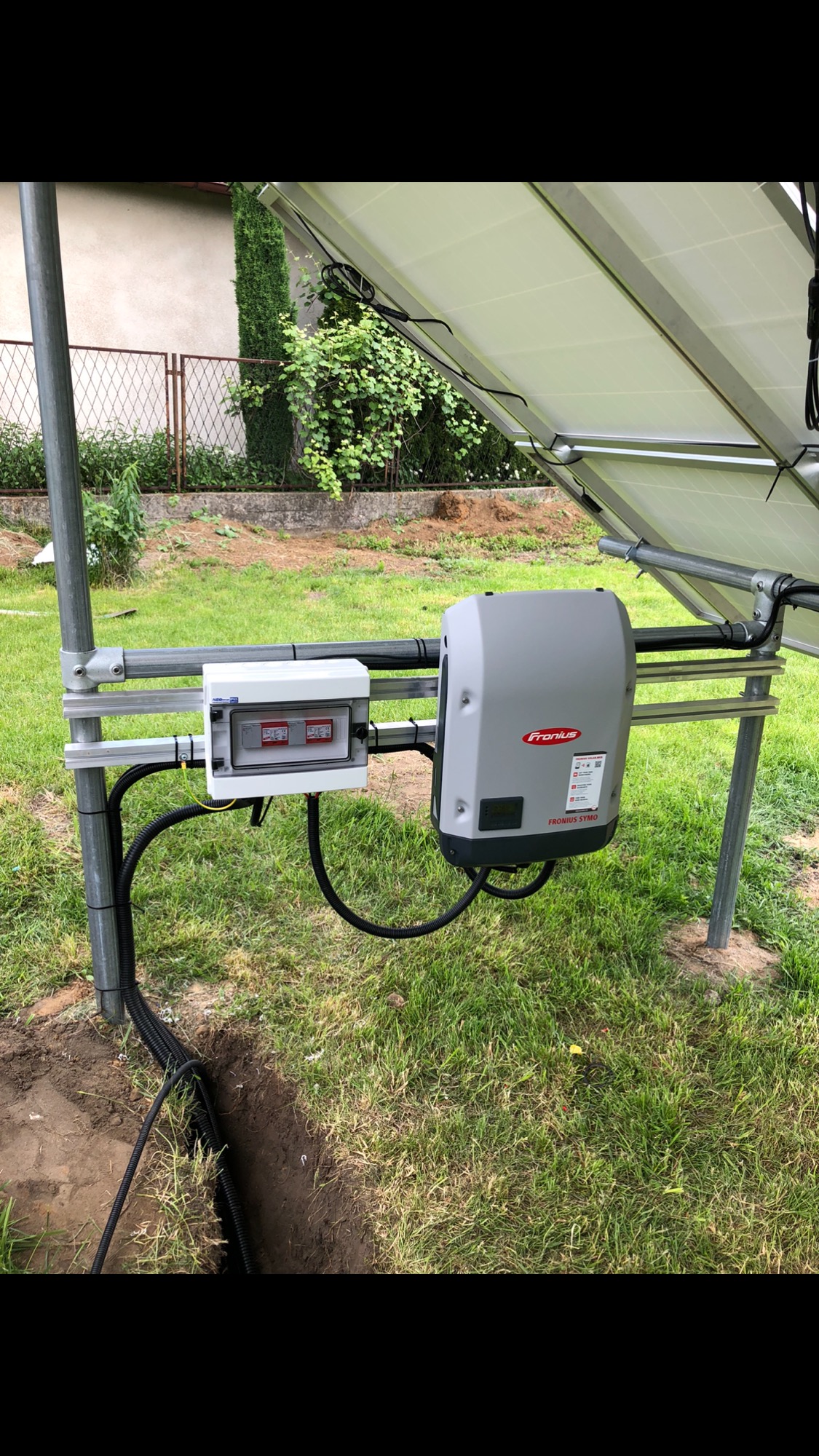 Montaż instalacji: Inwerter Fronius Symo i skrzynka zabezpieczeń przymocowane do konstrukcji wsporczej paneli słonecznych, widoczne kable prowadzone w ziemi.