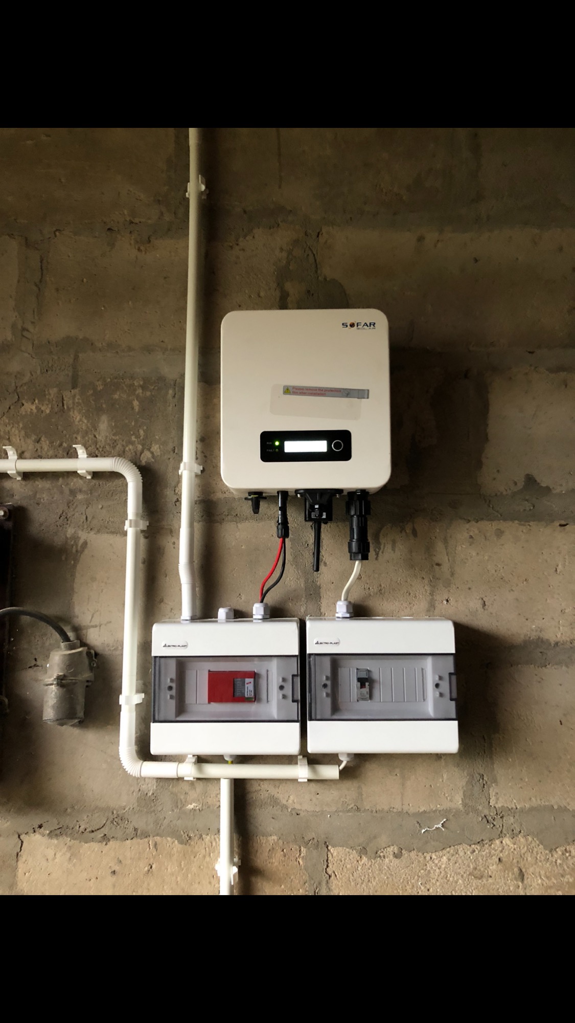 Zamontowany inwerter SOFAR z zabezpieczeniami elektrycznymi na surowej, betonowej ścianie. Widoczne przewody i rury instalacyjne.