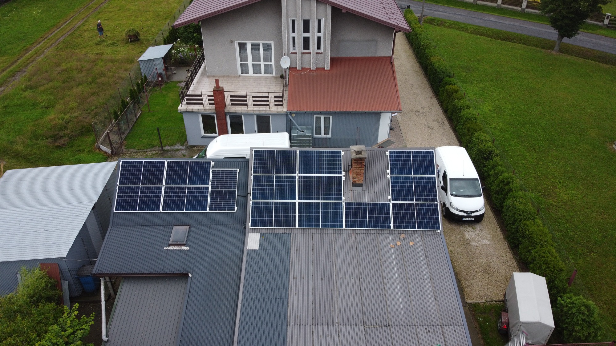 Widok z góry na instalację paneli słonecznych na dachu budynku gospodarczego, obok zaparkowany biały samochód dostawczy, w tle dom jednorodzinny z czerwoną dachówką i zadbany trawnik.