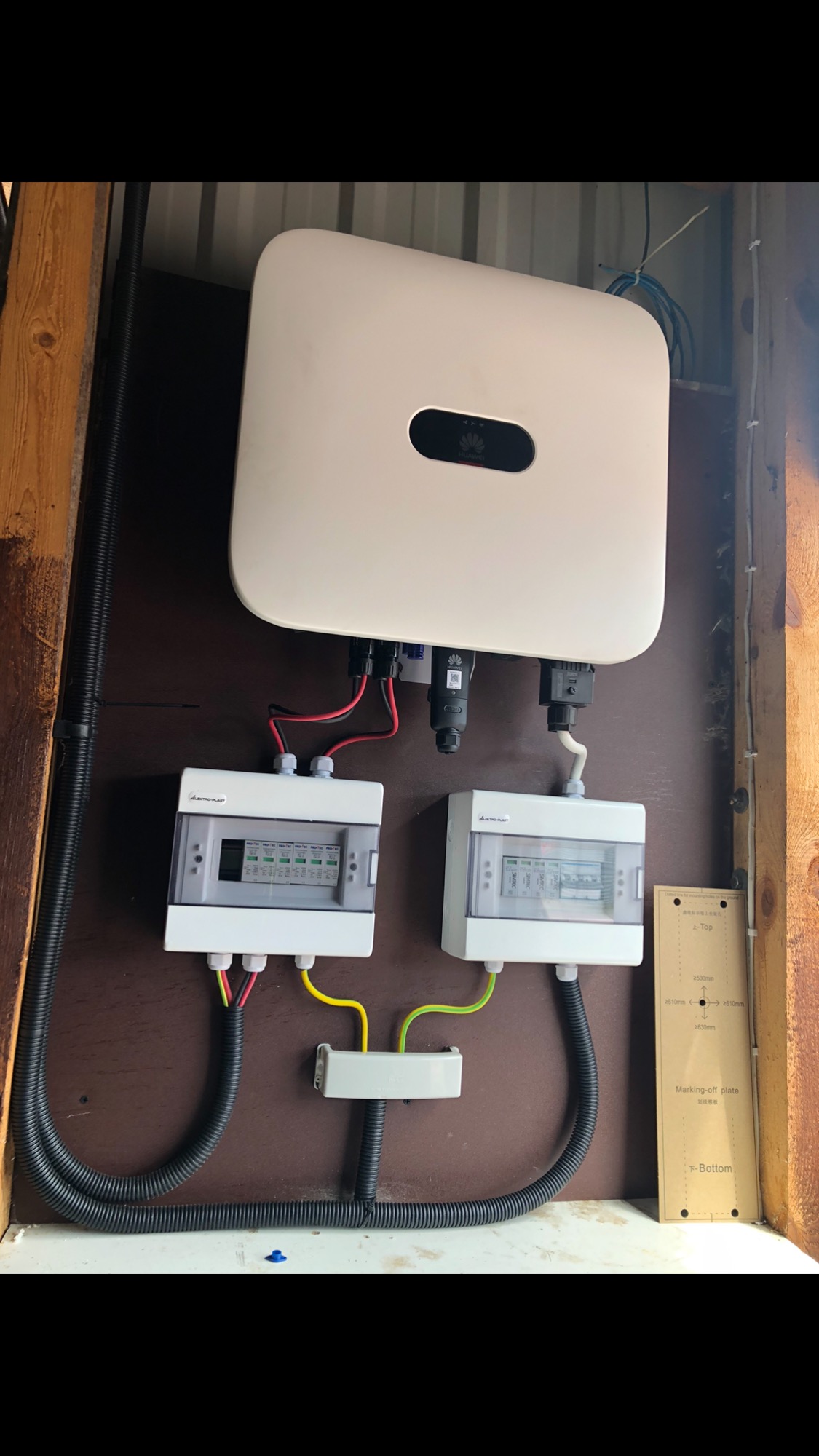 Biały falownik Huawei zamontowany na brązowej płycie wewnątrz pomieszczenia, z widocznymi dwoma skrzynkami bezpiecznikowymi i czarnymi rurami karbowanymi z kablami. Na ścianie obok skrzynki montażowe.