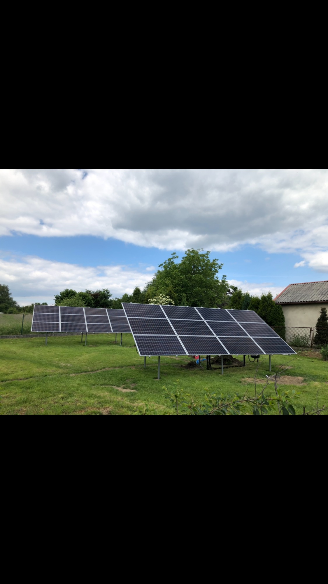 Rząd paneli słonecznych zamontowanych na trawniku przed domem jednorodzinnym, widoczne stalowe podpory i moduły fotowoltaiczne pod pochmurnym niebem.