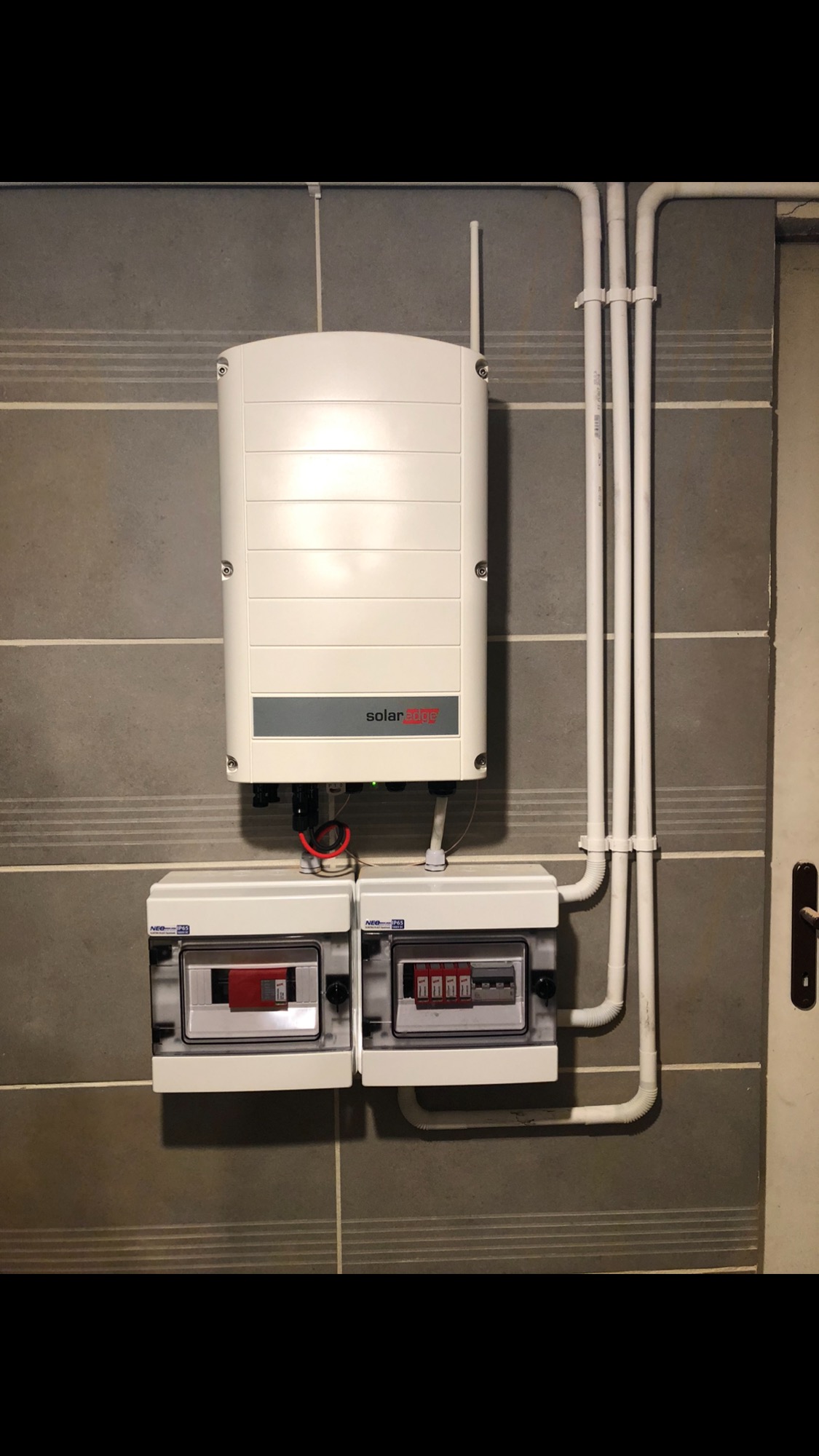 Biała obudowa falownika SolarEdge z anteną, zamontowana na szarej płytkowej ścianie nad dwiema skrzynkami zabezpieczeń, z widocznymi przewodami instalacji elektrycznej.