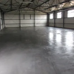 Wnętrze hali produkcyjnej z nową, szarą posadzką przemysłową, widoczne metalowe elementy konstrukcyjne budynku i okna w górnej części ścian.