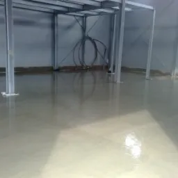 Świeżo wylana, gładka posadzka w hali przemysłowej, z widocznymi stalowymi słupami konstrukcyjnymi i zwiniętymi kablami pod sufitem.