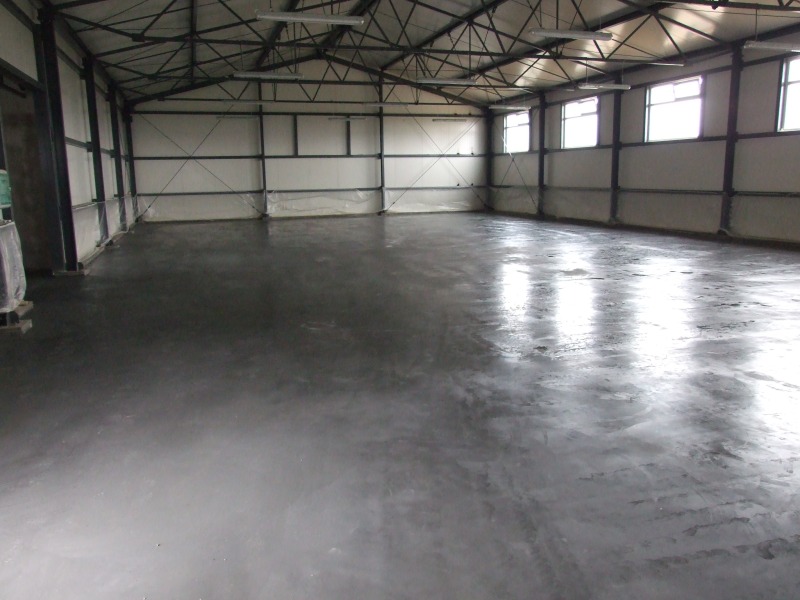 Wnętrze hali produkcyjnej z nową, szarą posadzką przemysłową, widoczne metalowe elementy konstrukcyjne budynku i okna w górnej części ścian.