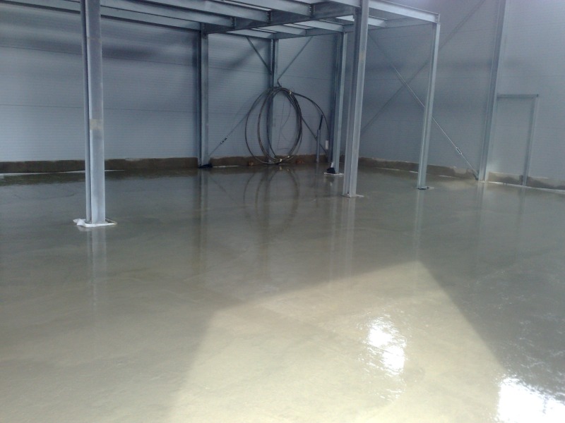 Świeżo wylana, gładka posadzka w hali przemysłowej, z widocznymi stalowymi słupami konstrukcyjnymi i zwiniętymi kablami pod sufitem.