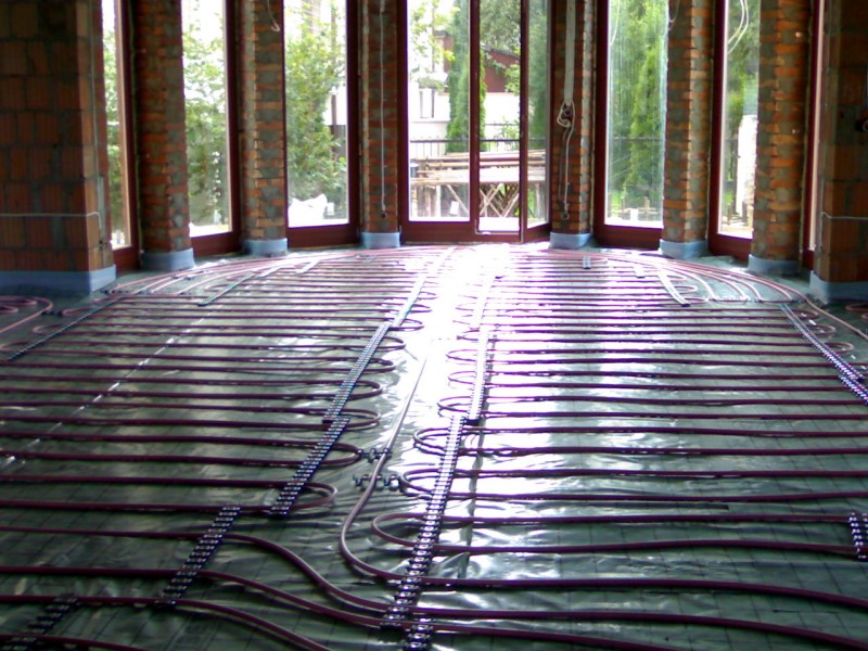 Rozłożona instalacja ogrzewania podłogowego w nowo budowanym pomieszczeniu z dużymi oknami i ceglanymi filarami, przygotowana do zalania wylewką.