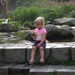 Wiktoria Hryncyszyn - Dziewczynka w różowej koszulce i granatowych spodenkach siedzi na kamiennym murku nad strumieniem, trzymając pluszowe zabawki.