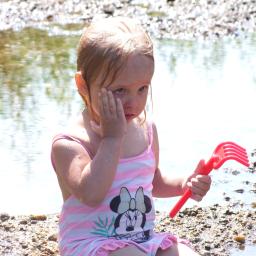 Wiktoria Hryncyszyn - Mała dziewczynka w różowo-białym stroju kąpielowym z motywem Myszki Minnie, siedząca na kamienistej plaży, trzymająca czerwoną zabawkę-grabki i zasłaniająca twarz dłonią, z wodą w tle.