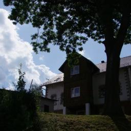 Wiktoria Hryncyszyn - Dwupiętrowy dom z ciemnego drewna z balkonem i białym budynkiem obok, częściowo zasłonięty przez drzewo i zieleń na pierwszym planie, pod błękitnym niebem z obłokami.