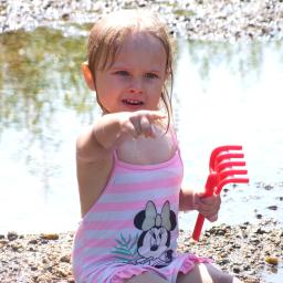 Wiktoria Hryncyszyn - Dziewczynka w stroju kąpielowym w paski z motywem Myszki Minnie, bawiąca się czerwoną grabką na plaży w Żaganiu, wskazuje palcem w stronę wody.