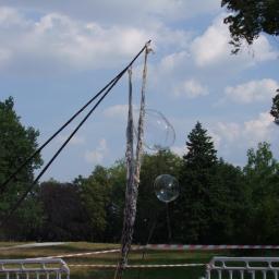 Wiktoria Hryncyszyn - Dwa duże bańki mydlane zawieszone w powietrzu, tworzone za pomocą specjalnego urządzenia z dwoma kijami i sznurkami, park w tle, słoneczny dzień.