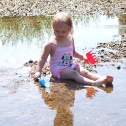 Wiktoria Hryncyszyn - Dziewczynka w stroju kąpielowym w paski z motywem Myszki Minnie siedzi w płytkiej wodzie na kamienistym brzegu, trzymając w rękach niebieską łopatkę i czerwoną grabkę.