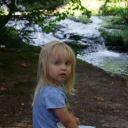 Dziewczynka z blond włosami i niebieskimi oczami siedzi na tle strumienia z małym wodospadem w lesie.