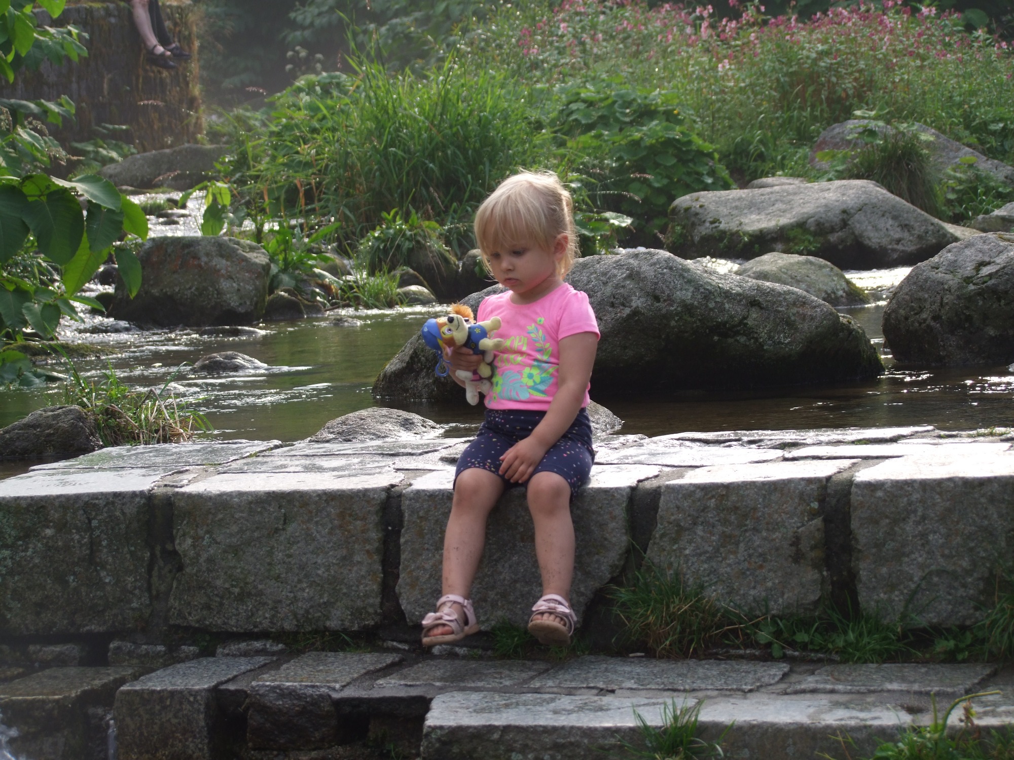 Dziewczynka w różowej koszulce i granatowych spodenkach siedzi na kamiennym murku nad strumieniem, trzymając pluszowe zabawki.