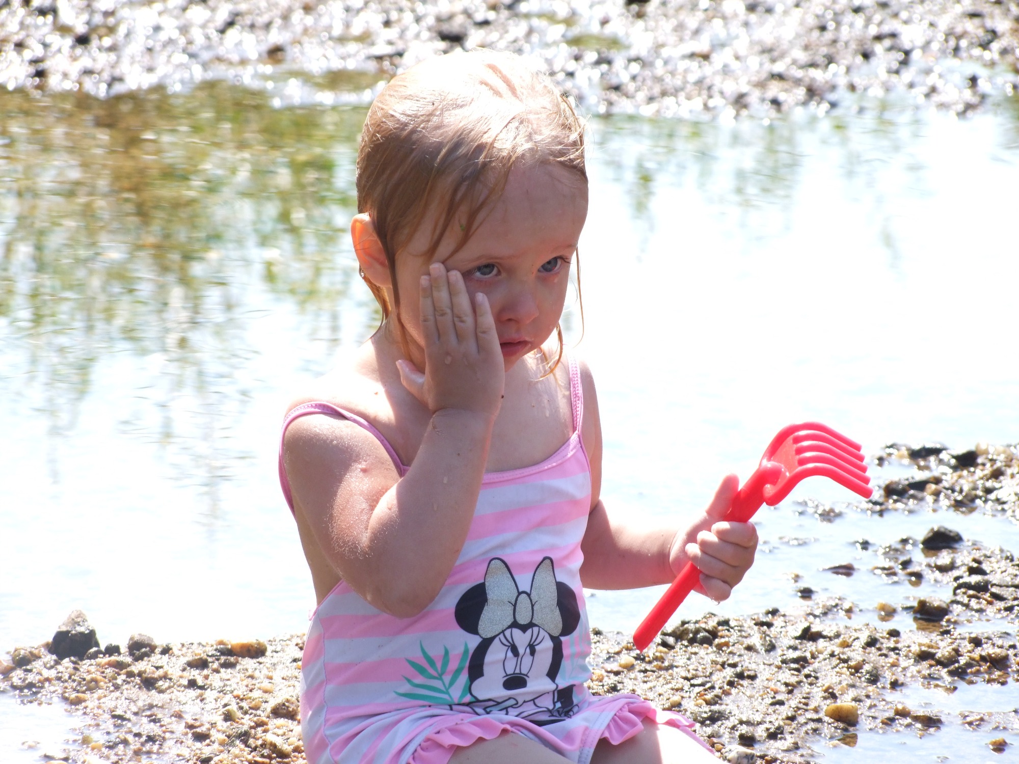 Mała dziewczynka w różowo-białym stroju kąpielowym z motywem Myszki Minnie, siedząca na kamienistej plaży, trzymająca czerwoną zabawkę-grabki i zasłaniająca twarz dłonią, z wodą w tle.