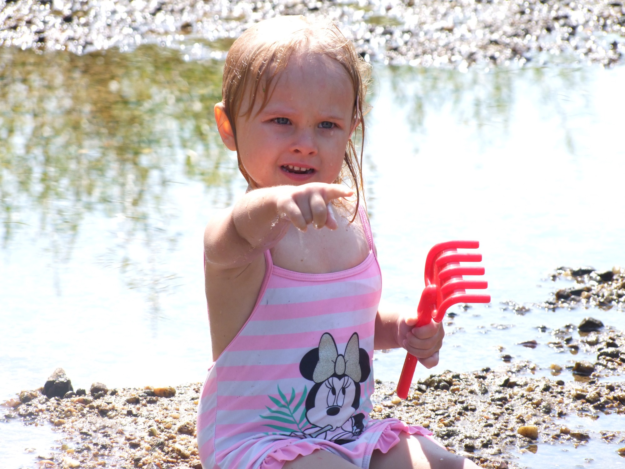 Dziewczynka w stroju kąpielowym w paski z motywem Myszki Minnie, bawiąca się czerwoną grabką na plaży w Żaganiu, wskazuje palcem w stronę wody.