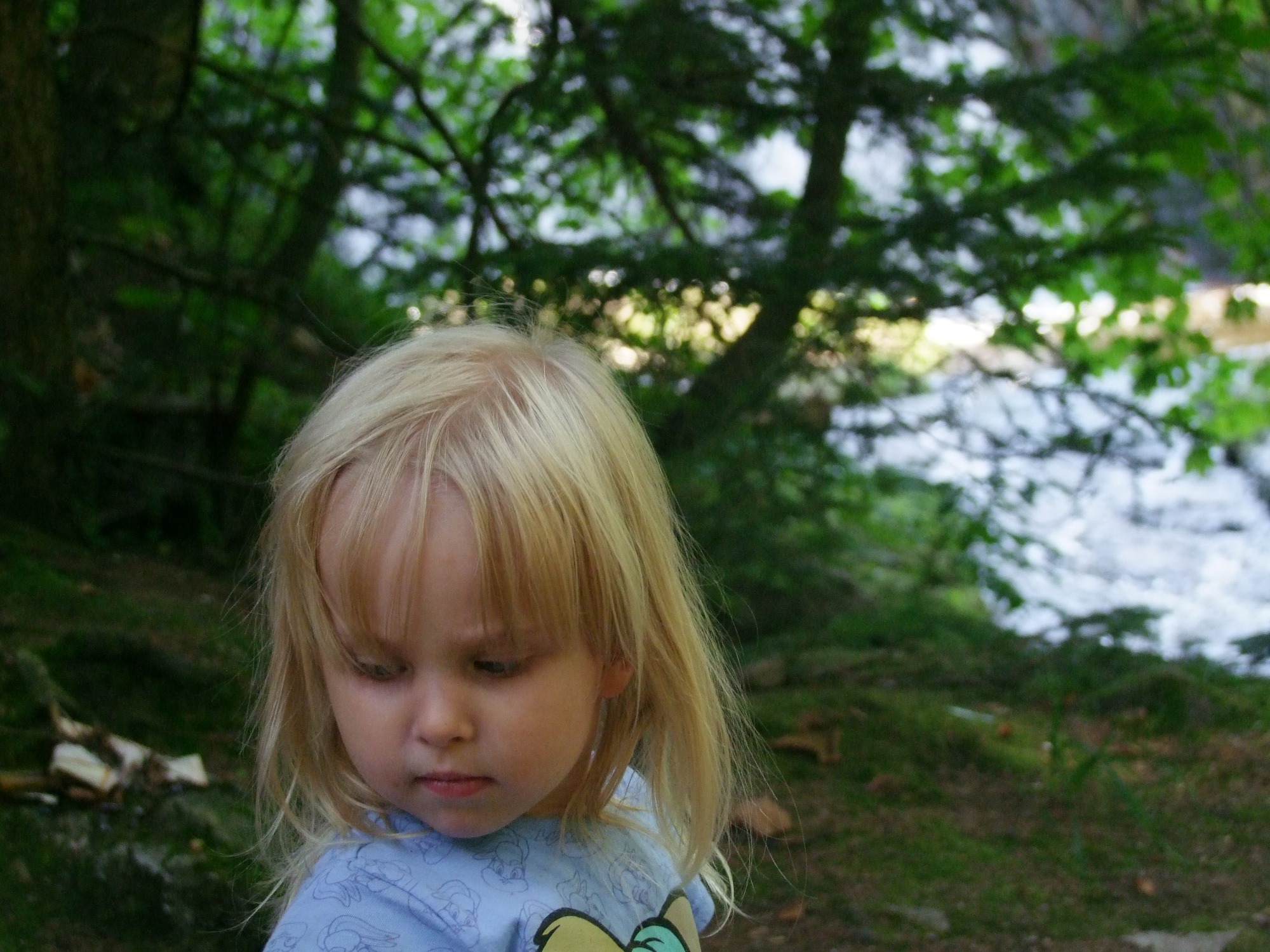 Portret małej dziewczynki z blond włosami na tle leśnego strumienia, zamyślony wyraz twarzy.