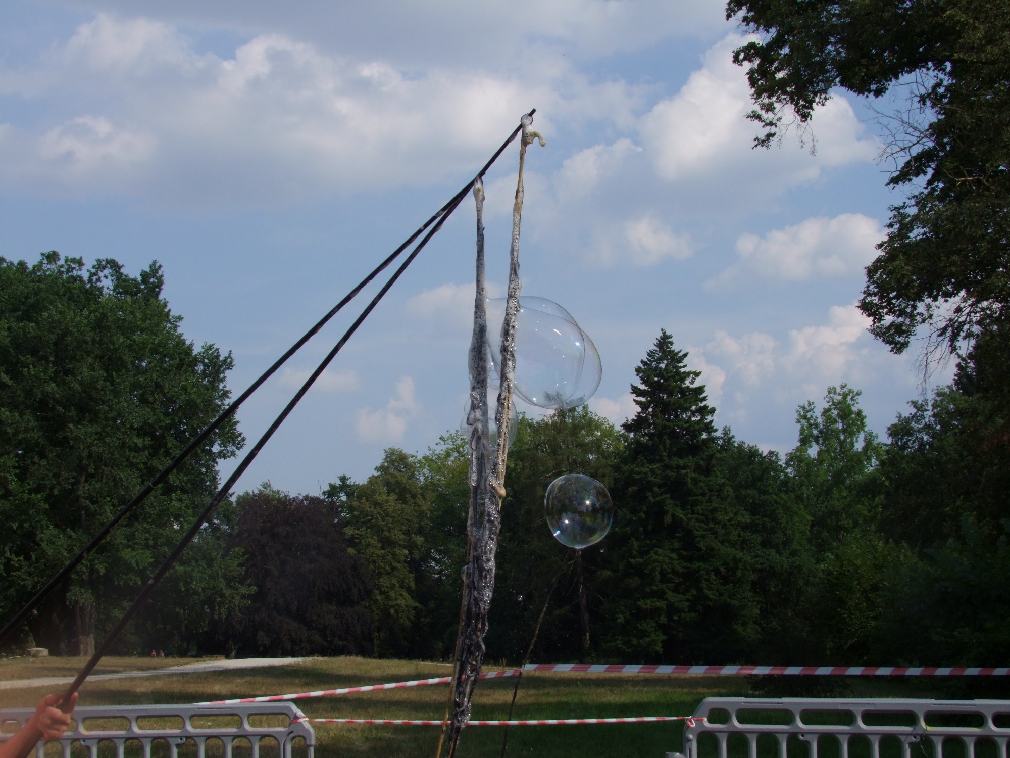 Dwa duże bańki mydlane zawieszone w powietrzu, tworzone za pomocą specjalnego urządzenia z dwoma kijami i sznurkami, park w tle, słoneczny dzień.