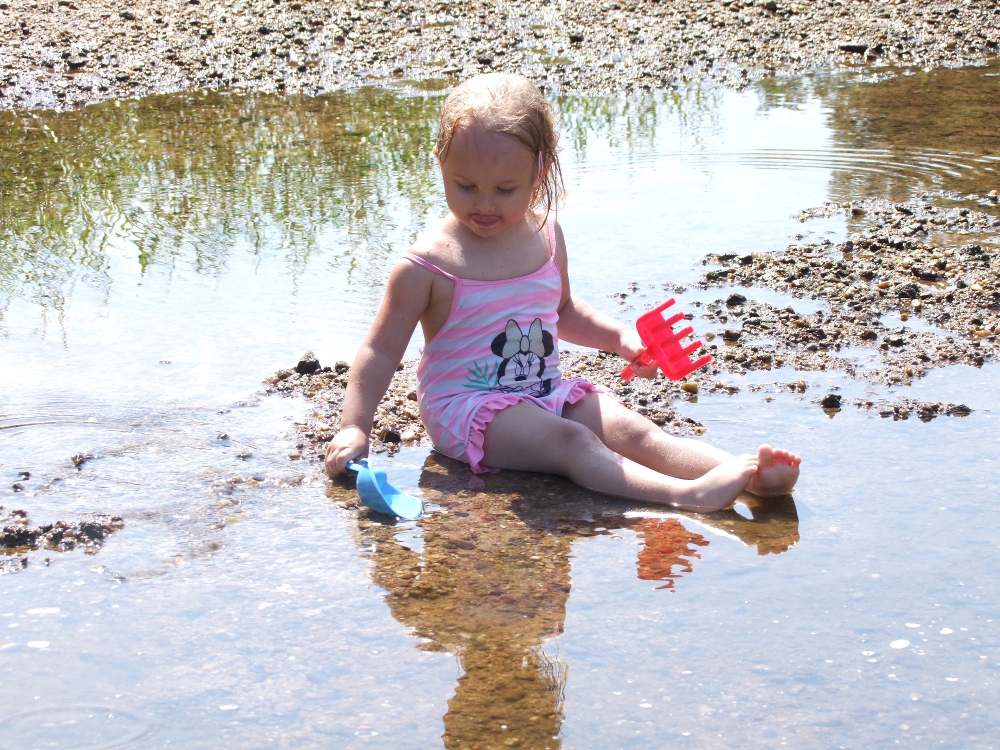 Dziewczynka w stroju kąpielowym w paski z motywem Myszki Minnie siedzi w płytkiej wodzie na kamienistym brzegu, trzymając w rękach niebieską łopatkę i czerwoną grabkę.