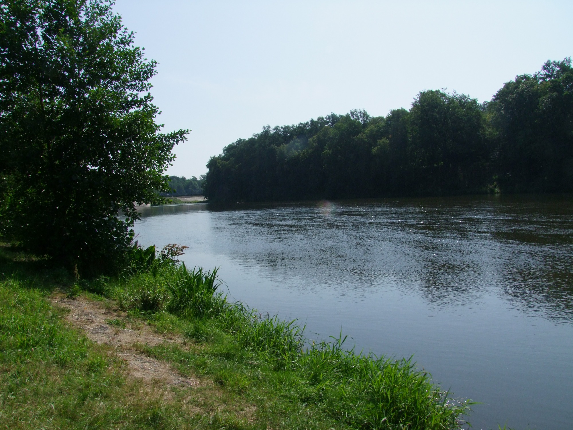 Spokojna rzeka otoczona zielenią drzew i traw, widoczna ścieżka wzdłuż brzegu.