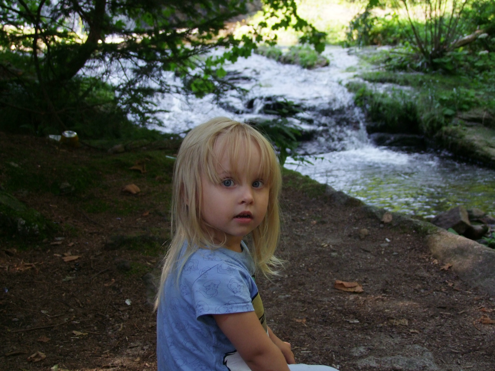 Dziewczynka z blond włosami i niebieskimi oczami siedzi na tle strumienia z małym wodospadem w lesie.