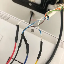 Fragment instalacji domofonu z widocznymi przewodami w kolorach czerwonym, czarnym, niebieskim i żółtym, zabezpieczonymi czarną taśmą izolacyjną, oraz otworem w ścianie.