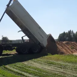 Ciężarówka z wywrotką wysypuje piasek na zieloną trawę, w tle widoczny las i niebo.